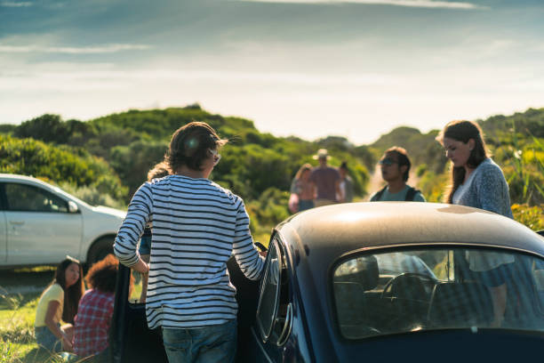 A photo of young friends standing by car. Men and woman are on road trip. They are in casuals on sunny day.