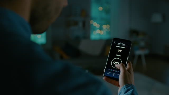 Close Up Shot of a Smartphone with Active Smart Home Application. Person is Tapping the Screen and Light is Being Turned On in the Room. It's Cozy Evening in the Apartment.