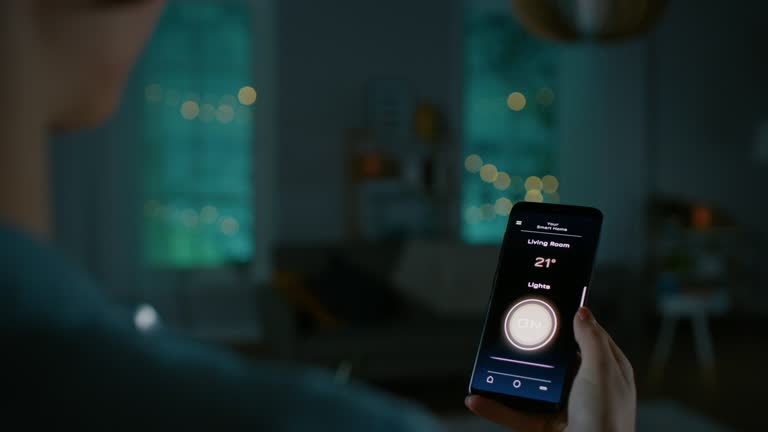 Young Beautiful Woman Gives a Voice Command to a Smart Home Application on Her Smartphone and Lights in the Room are Being Turned On. She Walks and Sits on a Couch. It's a Cozy Evening.