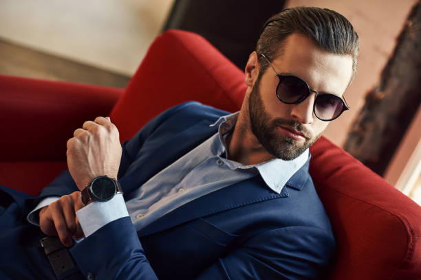 Looking just perfect...Young and sexy bearded businessman in sunglasses is looking away while sitting on office sofa Looking just perfect...Young and sexy bearded businessman in sunglasses is looking away while sitting on office sofa. Close-up. Business look. metrosexual stock pictures, royalty-free photos & images