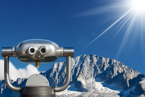 Coin operated electronic binoculars for tourists on a blurred landscape, mountain peak with glacier and blue sky with sun rays