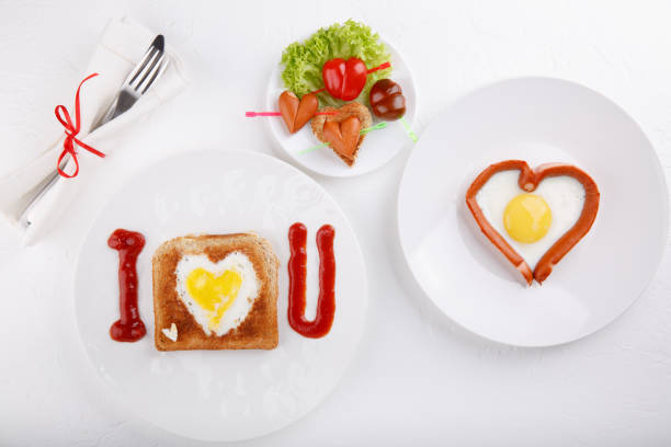 istock Toasted bread with fried eggs within, fried egg & hotdog on white plates. Heart Eggs. Festive breakfast on white table. Happy Valentines day. Top view.  Copy space. 1127490833