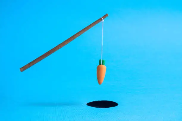 Photo of Orange carrot hanging on stick above black hole abstract isolated on blue.