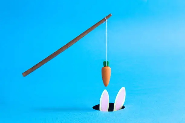 Photo of Orange carrot hanging on stick above black hole with rabbit in it abstract isolated on blue.