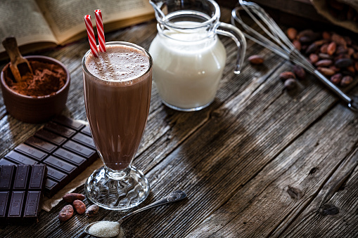 Preparing homemade chocolate milkshake. Chocolate milkshake glass shot on rustic wooden table. Some chocolate bars are beside the milkshake glass. A brown bowl filled with ground cocoa is at the left border of the frame. A milk pitcher is out of focus at background. An open cookbook is at the top left. The composition is at the left of an horizontal frame leaving useful copy space for text and/or logo at the right. Predominant color is brown. Low key DSRL studio photo taken with Canon EOS 5D Mk II and Canon EF 70-200mm f/2.8L IS II USM Telephoto Zoom Lens