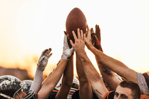 Football team holding the ball all together Group of men, American football team on training outdoors, holding hands all together. football helmet and ball stock pictures, royalty-free photos & images