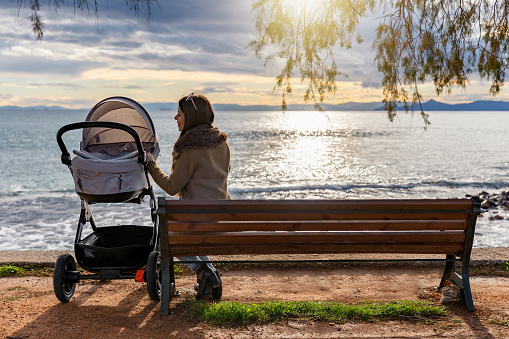 Mother sits on a bench in a park by the sea with her baby in a stroller and enjoys the sunset