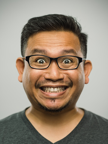 Close up portrait of asian man with very excited expression against white gray background. Vertical shot of malaysian real people surprised in studio with black hair and glasses. Photography from a DSLR camera. Sharp focus on eyes.