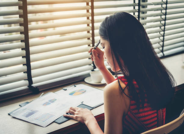 молодая женщина анализирует графики, сидя на столе в офисе - paper document pen long hair стоковые фото и изображения