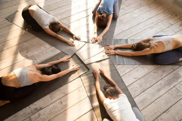 Fit female yogi stretch on rubber mats practicing yoga in fitness studio together, toned women relax in child pose lying on floor in circle, group of girls train doing pilates in gym. Wellness concept