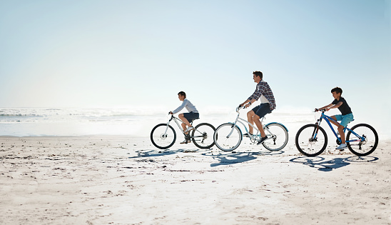 Shot of a father and his young sons riding bicycles together along the beach