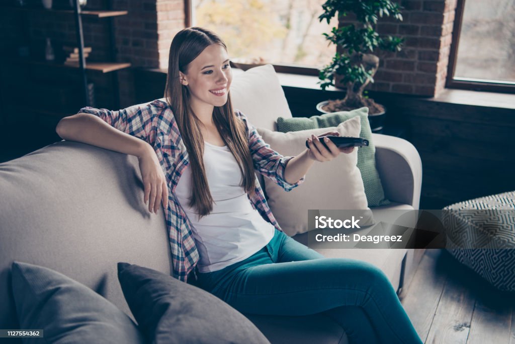 Attractive pretty cheerful glad positive optimistic cute lovely sweet charming she her lady using remote console for changing channels on tv lying on soft beige divan in light room Adult Stock Photo