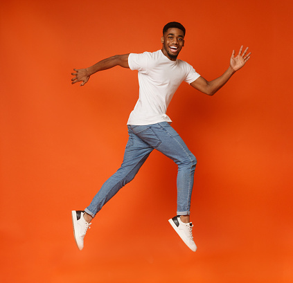 Funny young black millennial guy walking on air, jumping on orange studio background, smiling at camera