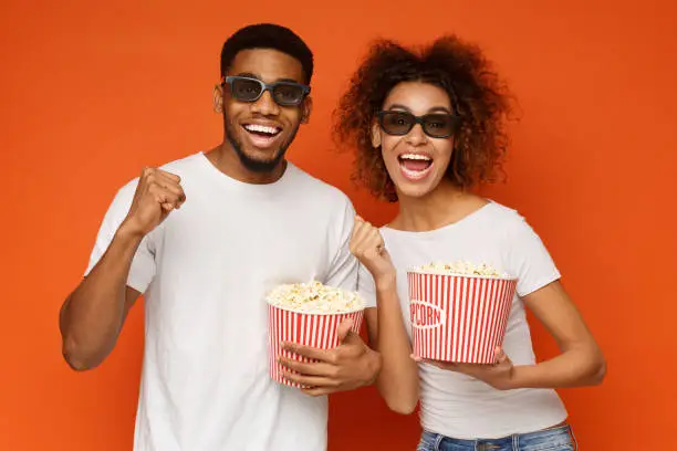 Enjoying modern innovative technology. Happy african-american couple in 3d glasses holding buckets of popcorn, orange background