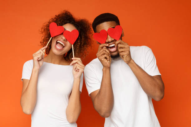 Young couple holding red love hearts over eyes Lovers blinded by their big love.. Young cheerful african-american couple in love holding red hearts over eyes and smiling, orange background male likeness photos stock pictures, royalty-free photos & images