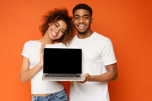 Young cheerful african-american couple in love holding laptop with blank screen, orange studio background