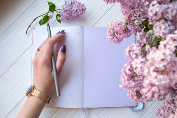 istock Lilac and notepad on a white desktop. Woman is writing in a notebook. Top view. 1127571155