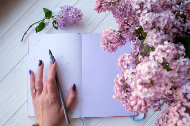 istock Lilac and notepad on a white desktop. Woman is writing in a notebook. Top view. 1127572286