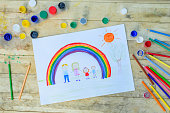 幸せな家族の概念。木製のテーブルの描画: 父、母、男の子と女の子は虹と晴れた空の背景に対して手を握る。
