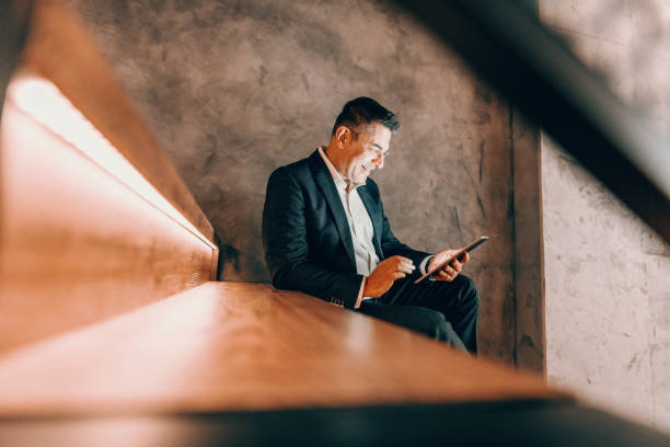 istock Middle aged man dressed smart casual using tablet for internet surfing and sitting on the stairs in cafeteria. 1127580524