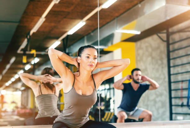 séduisante brunette faisant des squats dans la salle de gym. dans le miroir du fond et l’homme dans la réflexion. - crouching male men sensuality photos et images de collection