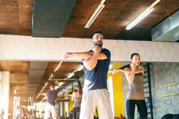 linda joven deportivo caucásica pareja en ropa deportiva estirando los brazos en el gimnasio. en el espejo del fondo. - stretching boyfriend indoors lifestyles fotografías e imágenes de stock