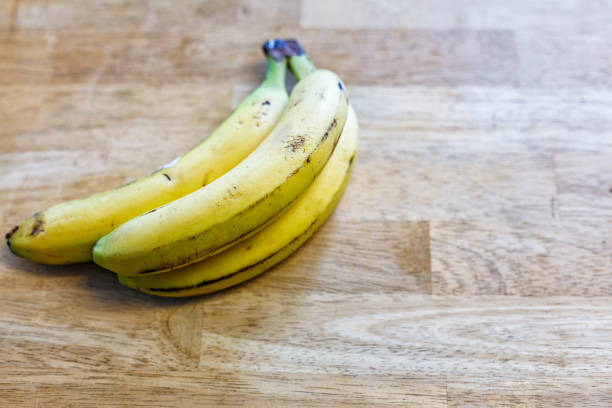 An unbroken, connected bunch of three ripe bananas still attached to each other on a kitchen wooden cutting board or table top surface. Potential concepts such as collaboration, connection, conformity, uniformity, linked, togetherness, groups, relationships, team work, passing time (as they ripen), etc.