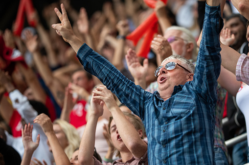 Portrait of a middle-aged man with sunglasses raising his arms in reaction to his team winning.