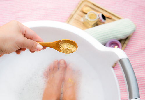 Woman taking a healing warming foot bath with mustard powder, adding mustard powder to foot bath with wooden spoon. Against cold illness, aches and improves blood circulation. Alternative medicine.