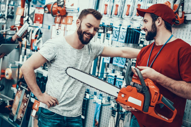 Salesman is Showing New Chainsaw to Client Salesman with Customer in Power Tools Store. Salesman is Bearded Caucasian Man. Customer is Caucasian Young Man. Seller is Showing New Chainsaw to Client. People is Happy and Smiling. hardware store stock pictures, royalty-free photos & images
