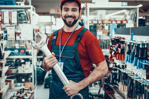 Salesman is Posing in Power Tools Store. Salesman is Young Bearded Caucasian Man. Man is Holding New Giant Wrench. Man is Happy and Smiling. Seller is Wearing Baseball Cap and T-Shirt.