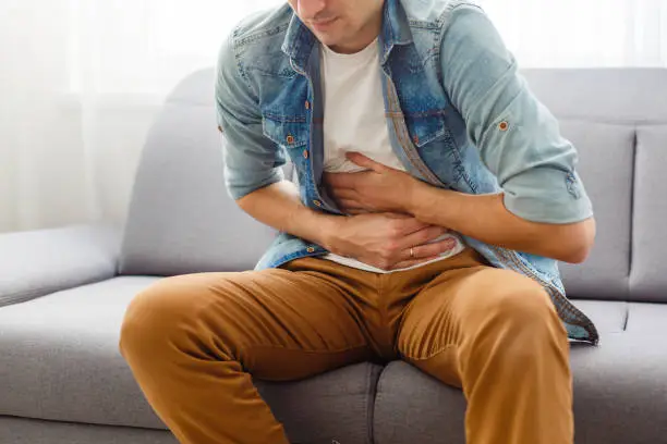 Photo of The man is sitting on a gray couch and holding his belly. Medicine and health concept, stomach problems. The man suffers from stomach ache, gastric problems. Abdominal pain, suffering and pain.
