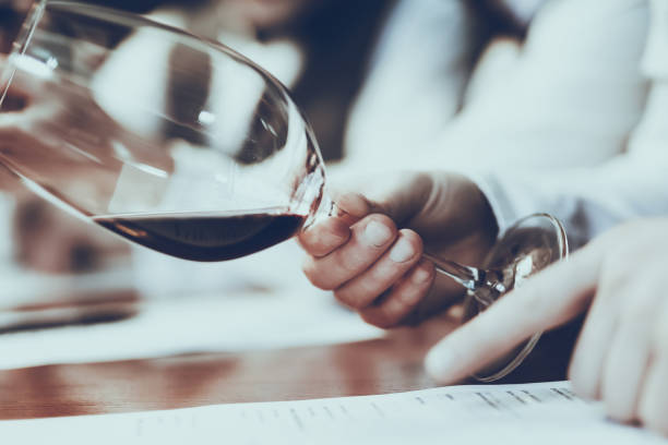 Experienced Sommelier is Working in Restaurant. Sommelier is Tasting Red Wine in Glass. Sommeliers is Man. Person is Sitting at Table. Man is Wearing Special Uniform. Closeup of Hand with Wine.