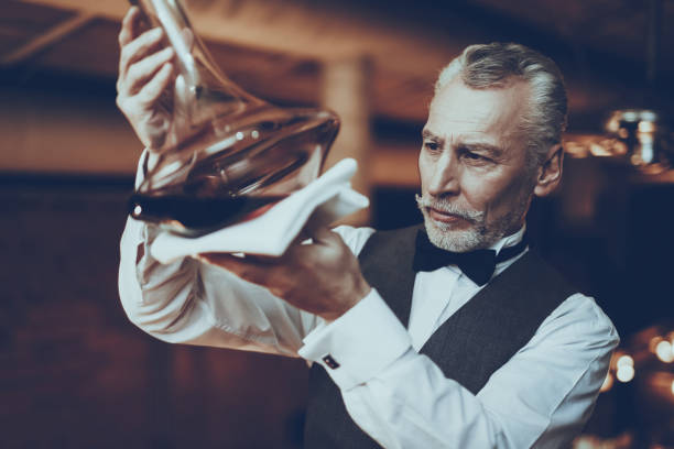 Sommelier Tasting Wine in Decanter in Restaurant Experienced Sommelier is Working in Restaurant. Sommelier is Looking on Wine Sediment in Decanter. Sommelier is Mature Caucasian Man. Person is Wears Special Uniform. Man is Degustates Wine. uncork wine stock pictures, royalty-free photos & images