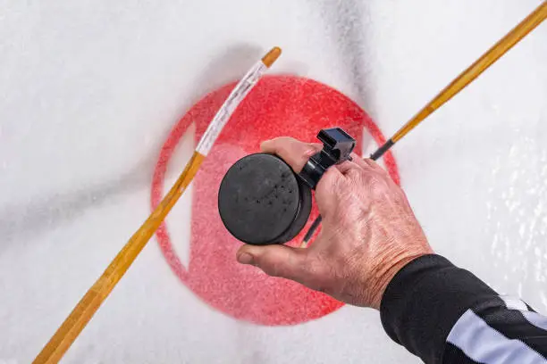 Looking down on a referee holding a black ice hockey puck over a red “Face-Off Spot” with two hockey sticks ready for the face-off.  The Face-Off Spot is used for players to faceoff as the ref drops the puck to resume play.