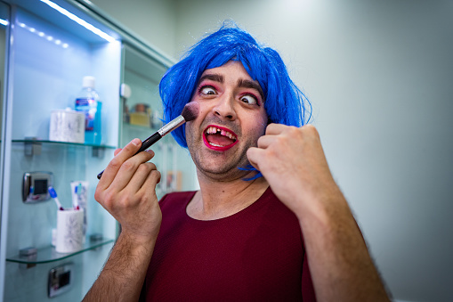 Toothless man, wearing blue wig, is putting on blush with make a up brush.