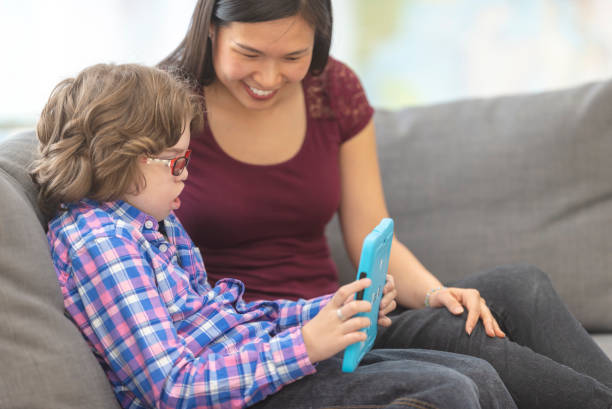 Student and his tutor using a tablet together A cute young boy who is deaf sits with his teacher of asian descent as they play on a tablet together while sitting on the couch. He is wearing glasses and has a tracheostomy. She is smiling while he plays a game. assistive technology stock pictures, royalty-free photos & images