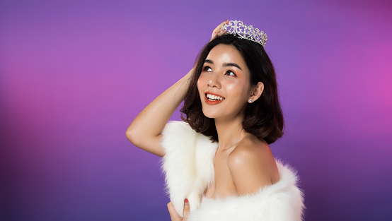 Portrait of Miss Pageant Beauty Contest in white fur Evening dress light Diamond Crown, LGBT transgender Asian Woman fashion make up black hair style, studio lighting gradient shade purple background