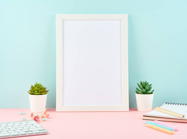 istock Mockup with blank white frame, alarm, notepad, keyboard on pink table against blue wall with copy space. Modern bright office desktop 1127690414