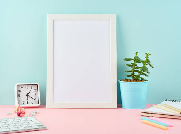 istock Mockup with blank white frame, alarm, notepad, cup of coffee on pink table against blue wall with copy space. 1127690418