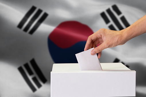 Close-up of human hand casting and inserting a vote and choosing and making a decision what he wants in polling box with South Korea flag blended in background