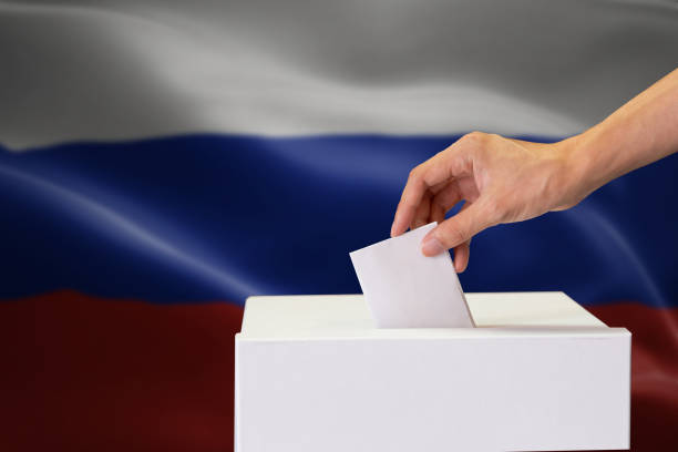 istock Close-up of human hand casting and inserting a vote and choosing and making a decision what he wants in polling box with Russia flag blended in background. 1127704164