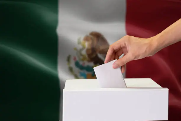 Close-up of human hand casting and inserting a vote and choosing and making a decision what he wants in polling box with Mexico flag blended in background
