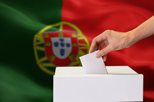 Close-up of human hand casting and inserting a vote and choosing and making a decision what he wants in polling box with Portugal flag blended in background