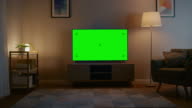 istock Zoom In Shot of a TV with Horizontal Green Screen Mock Up. Cozy Evening Living Room with a Chair and Lamps Turned On at Home. 1127715204