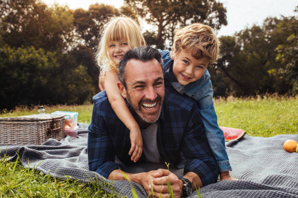 Children playing with their father on picnic Happy man with kids on a picnic lying down in park beside a picnic basket. Children lying on the back of their father. family with two children stock pictures, royalty-free photos & images