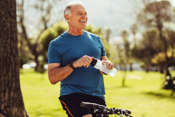 Senior fitness man drinking water Senior man in fitness wear drinking water sitting on his bicycle. Cheerful senior fitness person taking a break during cycling in a park. one senior man only stock pictures, royalty-free photos & images