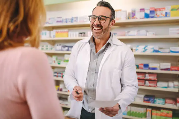 Helpful male pharmacist dealing with a woman customer standing at desk in the pharmacy. Smiling male pharmacist with prescription assisting a customer standing at the counter.