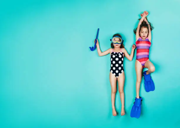 Two girls lying down with snorkelers and flippers. Top view of twin girls wearing swimwear on blue background.