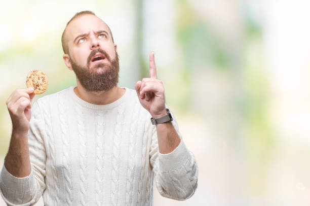 istock Young hipster man eating chocolate chips cookie over isolated background surprised with an idea or question pointing finger with happy face, number one 1127774121